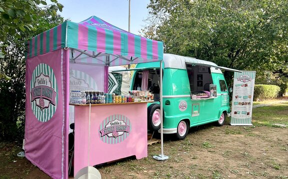 Sur l'image, on voit une tente pliante de 2x2 m, personnalisée, sur un pré, à côté d'un food truck de street food. La tente pliante a trois parois latérales imprimées en rose et bleu, tout comme le toit. Elle est fixée au sol avec des poids.