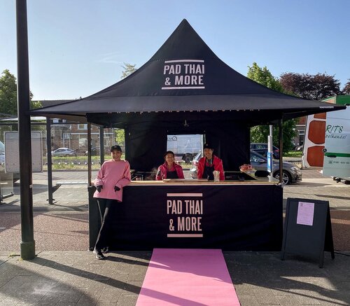 Auf dem Bild ist ein schwarzer 3x3 m Faltpavillon, bedruckt mit dem Logo von Pad Thai. Der Faltpavillon hat eine Seitenwand mit Tür und eine halbhohe Seitenwand mit Theke, wo das Essen serviert wird.