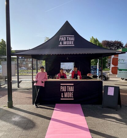 Auf dem Bild ist ein schwarzer 3x3 m Faltpavillon, bedruckt mit dem Logo von Pad Thai. Der Faltpavillon hat eine Seitenwand mit Tür und eine halbhohe Seitenwand mit Theke, wo das Essen serviert wird.