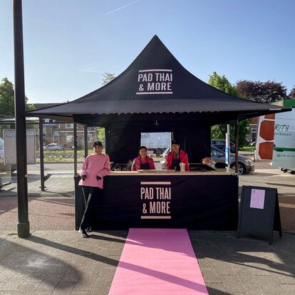 Nell'immagine c'è un gazebo pieghevole nero di 3x3 m, stampato con il logo di Pad Thai. Il gazebo pieghevole ha una parete laterale con porta e una parete laterale a media altura con un bancone dove viene servito il cibo.