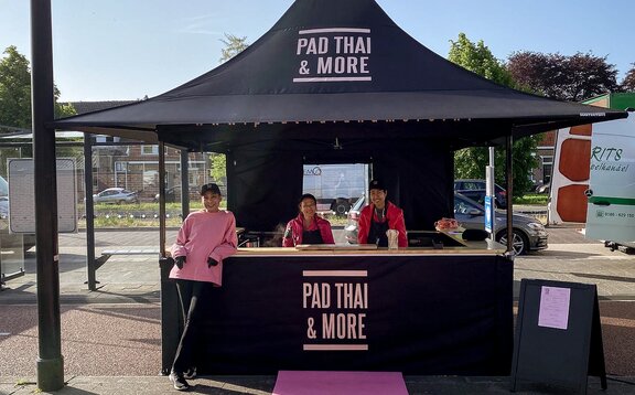 Sur l'image, on voit une tente pliante noire de 3x3 m, imprimée avec le logo de Pad Thai. La tente pliante a une paroi latérale avec une porte et une paroi latérale semi-haute avec un comptoir où la nourriture est servie.