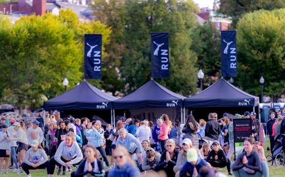 Três tendas dobráveis de 3x3 m estão dispostas lado a lado em um gramado durante um evento esportivo. As tendas dobráveis são pretas e personalizadas com o logo da Puma. Todas as três também possuem uma bandeira personalizada no teto. Muitas pessoas estão presentes.