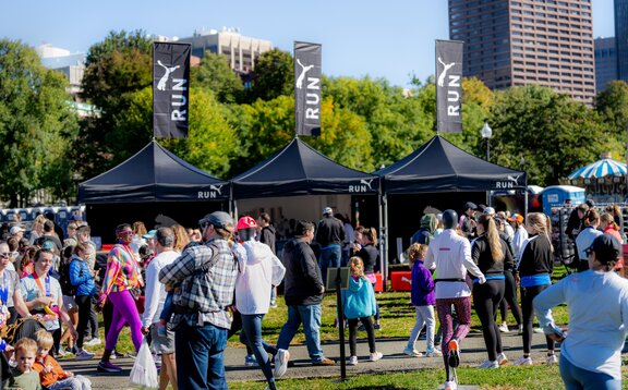 Tre gazebo pieghevoli da 3x3 m sono posizionati uno accanto all'altro su un prato durante un evento sportivo. I gazebo pieghevoli sono neri e personalizzati con il logo Puma. Tutti e tre hanno anche una bandiera personalizzata sul tetto. Ci sono molte persone presenti.