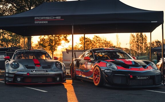 2 modelos Porsche Experience Racing uno al lado del otro bajo un pabellón negro plegable Oxford de 6x3 con panel recto y marco de aluminio negro al fondo de la puesta de sol.