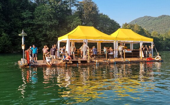 Rafting en un río de montaña con un grupo de turistas y 2 pabellones plegables Mastertent en amarillo y blanco hechos a medida para la protección contra la intemperie en una balsa con montañas al fondo.