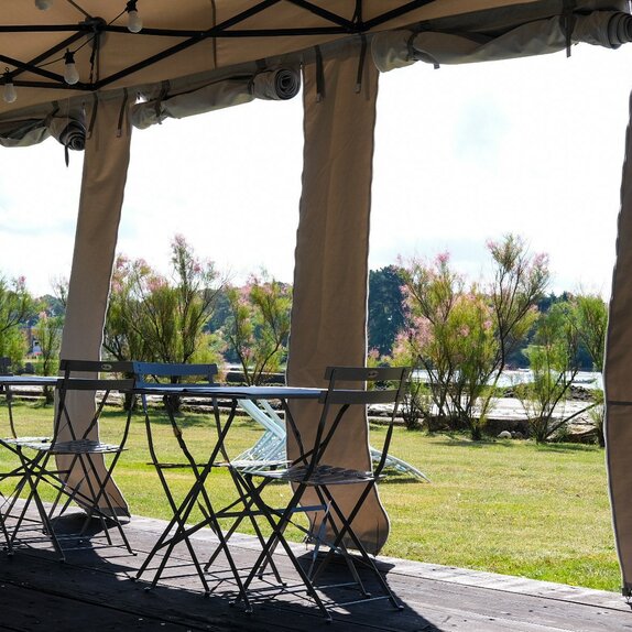 Inside to outside view of the expansive greenery through the custom-made open side panels of a white 6x4 folding pavilion with black aluminum construction on a wooden floor with beer garden seating and fairy lights