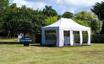 Folding pavilion 6x4 m in white with straight screen and side walls that can be rolled up according to customer requirements on a wooden terrace equipped with garden furniture and counter in a spacious green hotel complex with barbecue