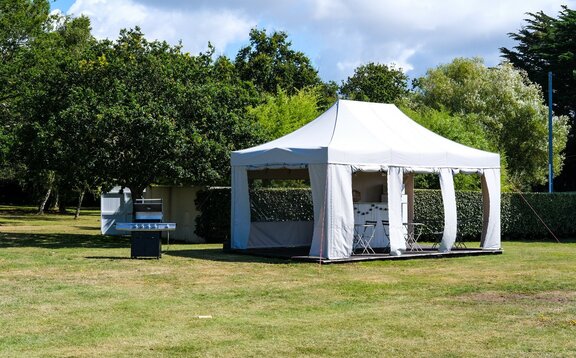 Faltpavillon 6x4 m in weiß mit gerader Blende und Seitenwänden nach Kundenwunsch nach oben aufrollbar auf einer Holzterrasse bestückt mit Gartenmobiliar und Tresen in einer weitläufigen grünen Hotelanlage mit Grill