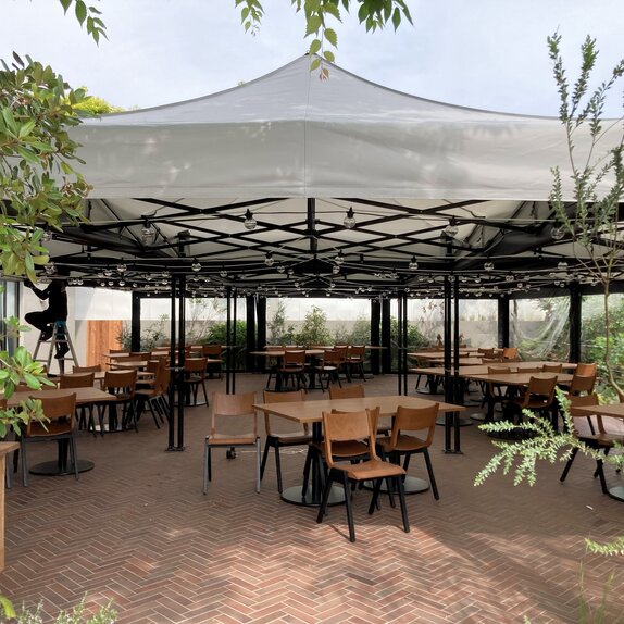 9 folding pavilions 4.5x3 m in a modular system on a stone terrace with gastronomic seating groups and green plants all around form a large tent with a white roof with a straight panel and black aluminum construction with specially manufactured side panels made of transparent PVC