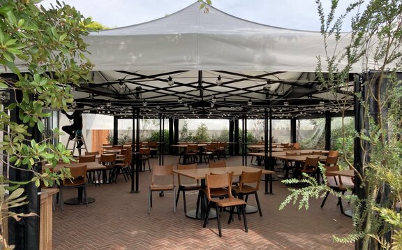 9 pabellones plegables de 4,5x3 m en sistema modular sobre una terraza de piedra con grupos de asientos gastronómicos y plantas verdes alrededor forman una gran carpa con techo blanco de panel recto y construcción de aluminio negro con paneles laterales especialmente fabricados de PVC transparente