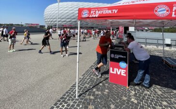 Folding pavilion 3x3 with white aluminum construction and personalized roof in red printed with FC Bayern Live TV logo with 2 promotion desks with laptops and employees talking to customers on the forecourt of the Allianz Arena with many spectators on their way to the stadium