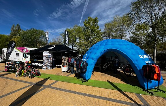 Decathlon promotion stand at open-air leisure fair with 4.5 x 3 m folding pavilion in black with side walls, counter half the length of the tent, 2 roof flags and beach flag as well as inflatable blue event tent model MS5 equipped with sales items such as bicycles and sportswear.