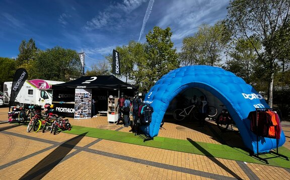 Stand de promotion de Decathlon sur un salon de loisirs en plein air avec un pavillon pliable de 4,5x3 m en noir avec des parois latérales, un comptoir à mi-longueur de la tente, 2 drapeaux de toit et un beachflag ainsi qu'une tente événementielle gonflable bleue modèle MS5 équipée d'articles de vente tels que des vélos et des vêtements de sport.