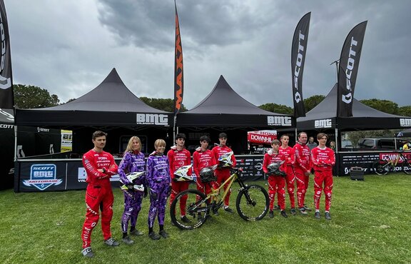 Three 5x5 m folding gazebos stand on a lawn. They are black and personalised with white logos from Scott, BMT and Honda. All three folding gazebos have half-height sidewalls and printed rear walls, as well as large, personalised flags on the roof. The racers are in the foreground.