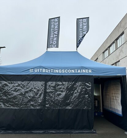 The photo shows a blue 6x4 m folding gazebo. It has two large windows at the front and a door through which you can see into the tent. You can see that the tent is printed on the inside of the right-hand side wall. It also has two flags on the roof. A white logo can be seen on the roof and both flags.
