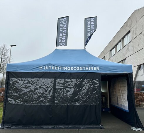 The photo shows a blue 6x4 m folding gazebo. It has two large windows at the front and a door through which you can see into the tent. You can see that the tent is printed on the inside of the right-hand side wall. It also has two flags on the roof. A white logo can be seen on the roof and both flags.