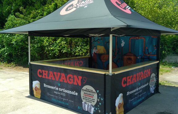 The photo shows a black 3x3 m folding gazebo printed with personalised motifs and a logo. The gazebo has 4 awnings and a counter on 3 sides. The rear wall is fully printed. Bushes and trees can be seen in the background.