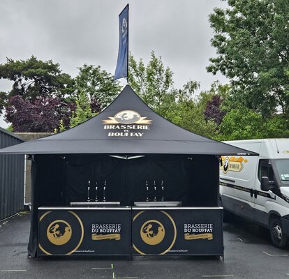 The picture shows a black 3x3 m folding gazebo with a personalised print on the sidewalls, roof and roof flag. The gazebo also has four awnings.