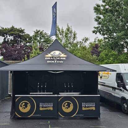 Es ist ein schwarzer 3x3 m Faltpavillon mit personalisiertem Druck auf den Seitenwänden, dem Dach und der Dachfahne zu sehen. Außerdem hat der Faltpavillon vier Vordächer.