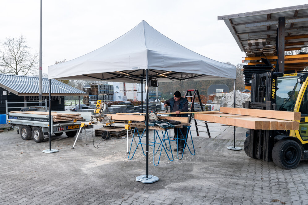 The picture shows a white 3x3 m folding gazebo that serves as a work tent. There is a person cutting wooden panels under the tent. The gazebo is secured to the ground with weights.