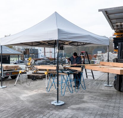 The picture shows a white 3x3 m folding gazebo that serves as a work tent. There is a person cutting wooden panels under the tent. The gazebo is secured to the ground with weights.