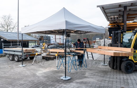 The picture shows a white 3x3 m folding gazebo that serves as a work tent. There is a person cutting wooden panels under the tent. The gazebo is secured to the ground with weights.