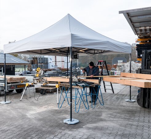 Auf dem Bild ist ein weißer Faltpavillon 3x3 m, der als Arbeitszelt dient. Unter dem Zelt befindet sich eine Person, die Holzplatten schneidet. Der Faltpavillon ist mit Gewichten am Boden befestigt.