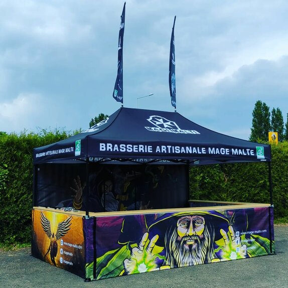 The picture shows a fully printed 4.5 x 3 m folding gazebo. It is black and its roof has the brewery's logo. Its rear wall and the three half-height side walls are printed all over with motifs of magicians. The folding gazebo has a counter on three sides and two flags on the roof.