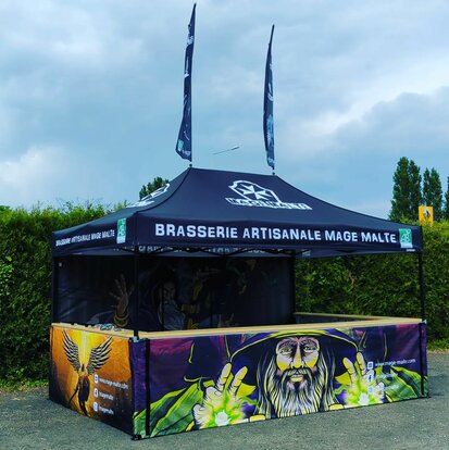Auf dem Bild ist ein vollflächig bedruckter Faltpavillon 4,5x3 m zu sehen. Er ist schwarz und sein Dach hat das Logo der Brauerei. Seine Rückwand sowie die drei halbhohen Seitenwände sind vollflächig mit Motiven von Zauberern bedruckt. Der Faltpavillon hat an drei Seiten eine Theke und zwei Flaggen auf dem Dach.