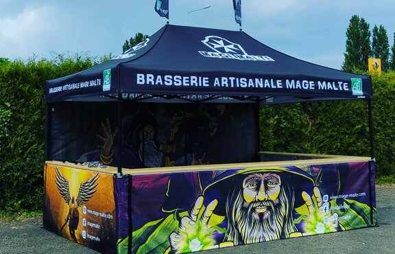 The picture shows a fully printed 4.5 x 3 m folding gazebo. It is black and its roof has the brewery's logo. Its rear wall and the three half-height side walls are printed all over with motifs of magicians. The folding gazebo has a counter on three sides and two flags on the roof.