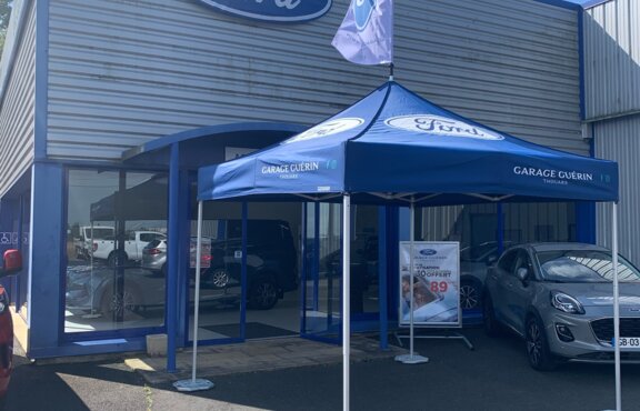 The photo shows a blue 3x3 m gazebo in front of a car dealership. The Ford logo can be seen on the roof of the gazebo and on its roof flag. The gazebo is attached to the ground with weights.