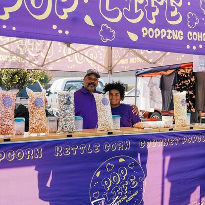 Tenda pieghevole viola con scritta gialla, sotto padre e figlio che vendono popcorn.