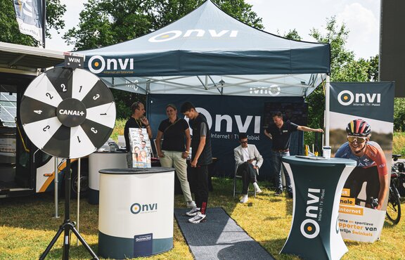 There is a dark blue 3x3 m folding gazebo on a meadow. The folding gazebo is printed with the Onvi logo, which can also be seen on the roof flag. The event tent has a side wall that is printed all over and a white structure. There are people under the tent and an advertising banner and tables in the foreground.