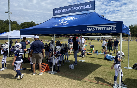 A blue folding gazebo is standing on a sports field. It has a printed banner and a printed roof. It is attached to the ground with weights. Several people are standing under the folding gazebo. 