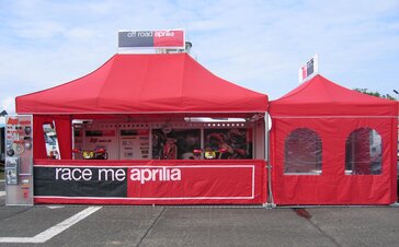 Ein roter Faltpavillon mit dem Schriftzug " Race me aprilia" steht neben einem kleineren Faltpavillon mit Seitenwand mit Fenstern. Unter dem großen Faltpavillon stehen zwei Motorräder