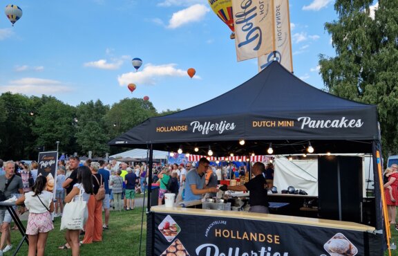 A black folding gazebo is standing on a field. It has a printed roof and is decorated. It has sidewalls and a counter. It has two printed flags and is secured to the ground with weights.