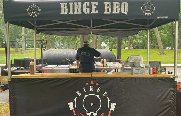 A black folding gazebo is standing in a park. It has a printed roof, a counter and a half-height sidewall. It is attached to the floor with weights.