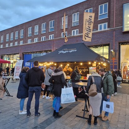 Ein schwarzer Faltpavillon steht auf einem Markt. Er hat ein bedrucktes Dach, Seitenwände und eine Theke.  Er hat zwei bedruckte Fahnen und ist mit Gewichten am Boden befestigt.