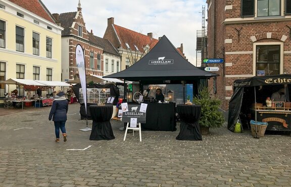 A black folding gazebo stands at a market. It has a printed roof and  sidewalls with windows. It is secured to the ground with weights.