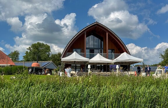 Three gray folding gazebos are standing next to a meadow in front of a building. Under them, there is a row of tables.