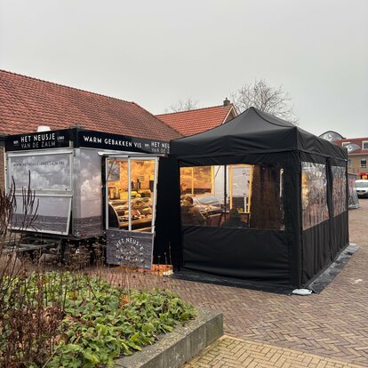Un gazebo pieghevole nero è fissato al suolo con dei pesi. Ha pareti laterali con finestre e una porta.