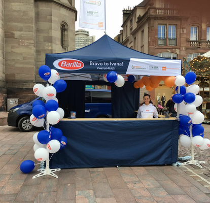 Ein blau-weißer Informationsstand der Firma "Barilla" ist mit Luftballons geschmückt. Darunter steht eine lachende Mitarbeiterin in einem weißen Pullover.