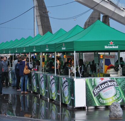 Getränkestände, bedruckt mit Bildern des Heineken Biers und dem Logo "Heineken", stehen auf einem Festivalgelände. Die Getränkestände sind gut besucht.