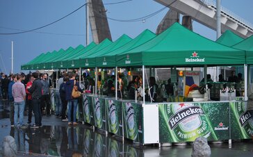 Standuri pentru băuturi, imprimate cu imagini ale berii Heineken și cu logo-ul "Heineken", se află la un festival. Standurile pentru băuturi sunt foarte frecventate.