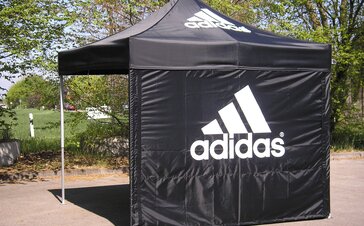 Ein schwarzer Faltpavillon mit einer geschlossenen Seitenwand, bedruckt mit dem Logo "Adidas", steht auf einem asphaltierten Boden.