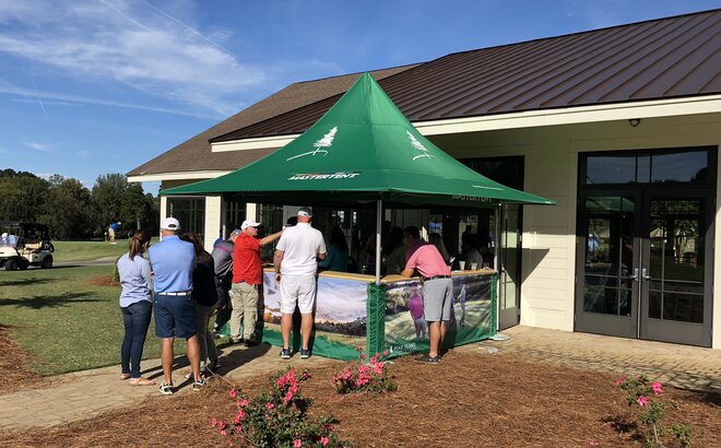Grüner Faltpavillon mit Vordach und bedruckten halbhohen Seitenwänden am Rande des Golfplatzes. Davor reihen sich Personen ein, die auf Informationen und Tickets warten.