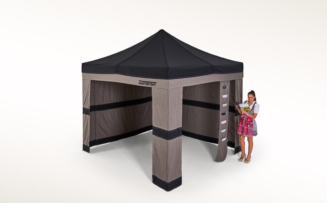Green Loden tent with two sidewalls at the back and a corner curtain at the front. Next to it a woman is standing in a traditional dress called Dirndl. 