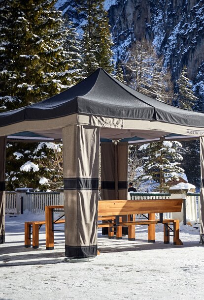 On a terrace there is a folding gazebo, underneath it a folding furniture. There is snow on the ground and on the trees and rocks in the background.