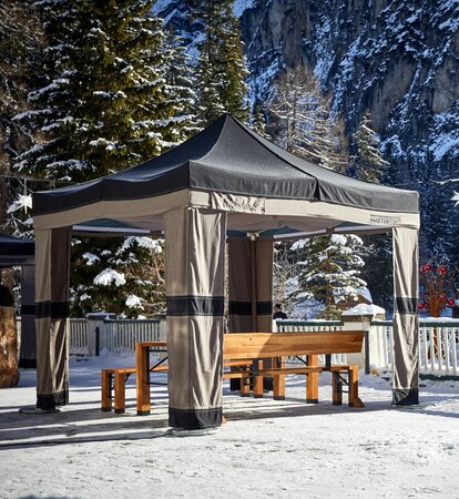 Su una terrazza c'è un gazebo pieghevole e sotto di esso un set di tavoli e panche richiudibili. Nello sfondo ci sono alberi e roccie innevati.