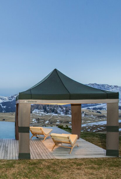 A Loden tent in the colours grey and green is located beside the pool at sunset. Under it are 2 wooden benches. 